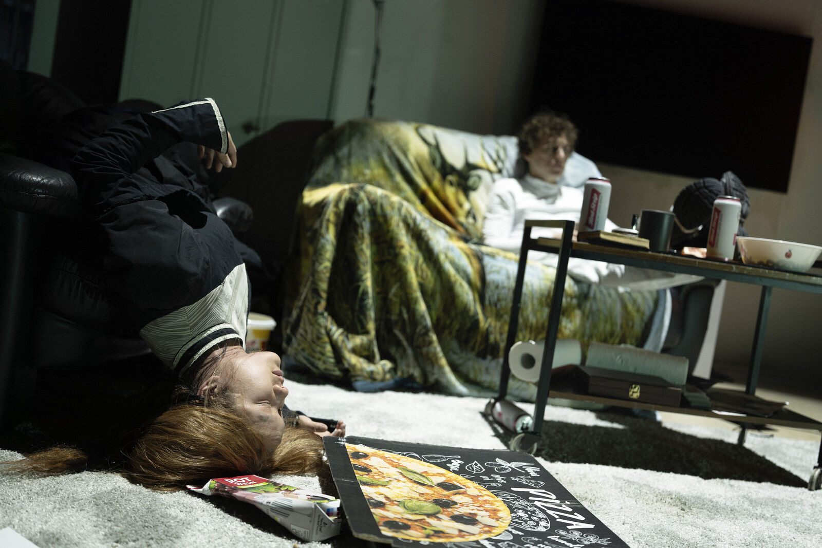 Eine Person liegt kopfüber auf einem Sofa. Auf dem Boden steht ein Pizzakarton. Eine andere Person sitzt, in eine Decke gehüllt, auf einem Sofa neben einem Tisch mit Getränkedosen. Der Raum ist schwach beleuchtet, was eine entspannte Atmosphäre schafft.