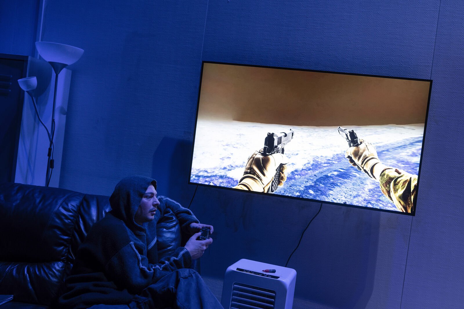 Eine Person in einem Kapuzenpullover spielt ein Videospiel auf einem großen Bildschirm. Das Spiel wird aus der Ich-Perspektive gezeigt. Der Raum ist schwach blau beleuchtet. Die Person sitzt mit einem Controller in der Hand auf einem Sofa. In der Nähe befinden sich eine Stehlampe und ein kleiner Heizstrahler.