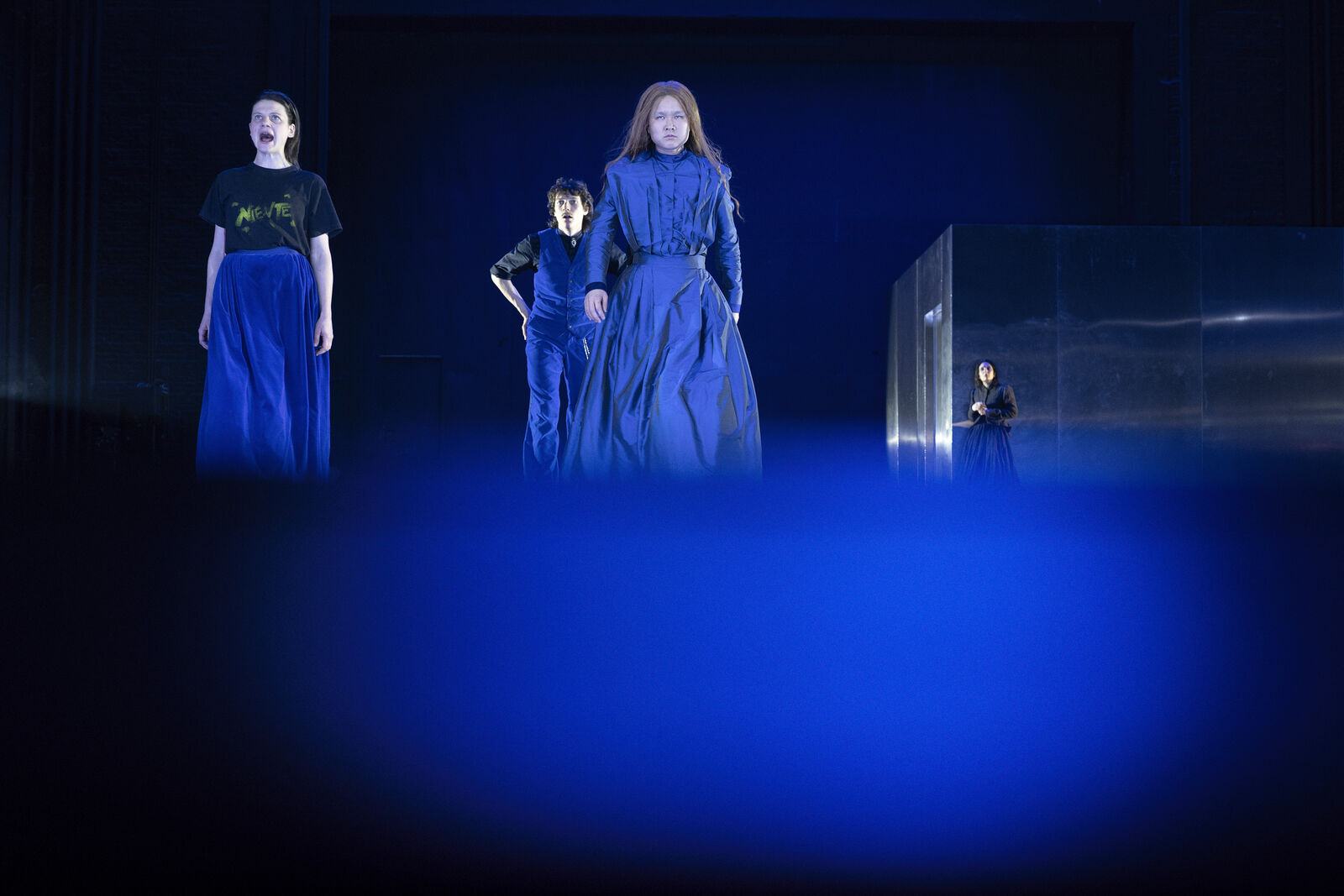 Vier Personen in einer Theaterszene mit blau beleuchteter Bühne. Sie tragen dunkle Kleidung, eine Person rechts ist durch reflektierende Elemente voneinander getrennt. Die Atmosphäre ist dramatisch und intensiv.