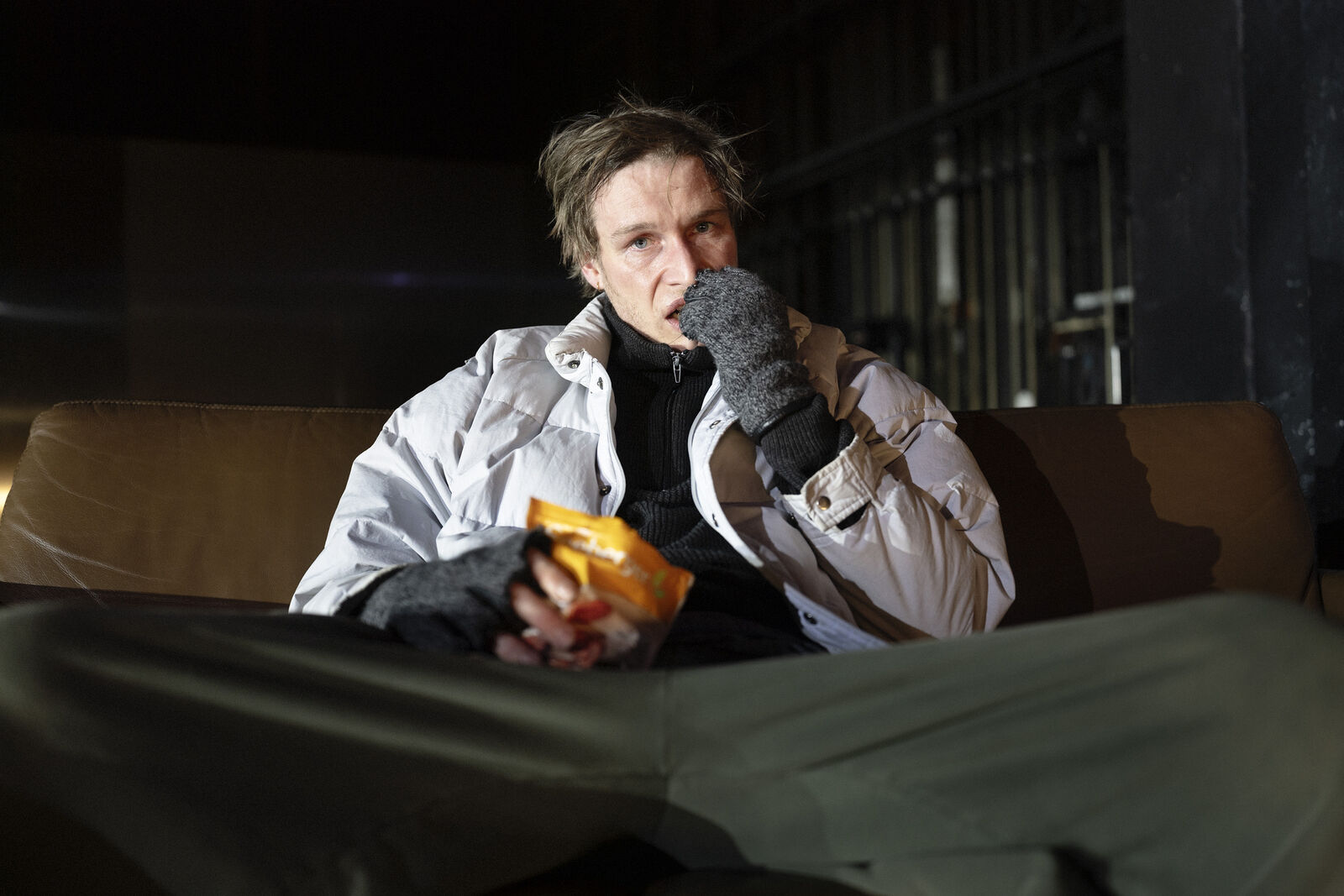 Eine Person sitzt in einem schwach beleuchteten Raum auf einem Sofa. Sie trägt eine weiße Jacke, graue Handschuhe und eine grüne Hose. Sie hält eine Tüte mit Snacks in der Hand und scheint mit nachdenklichem Gesichtsausdruck zu essen.