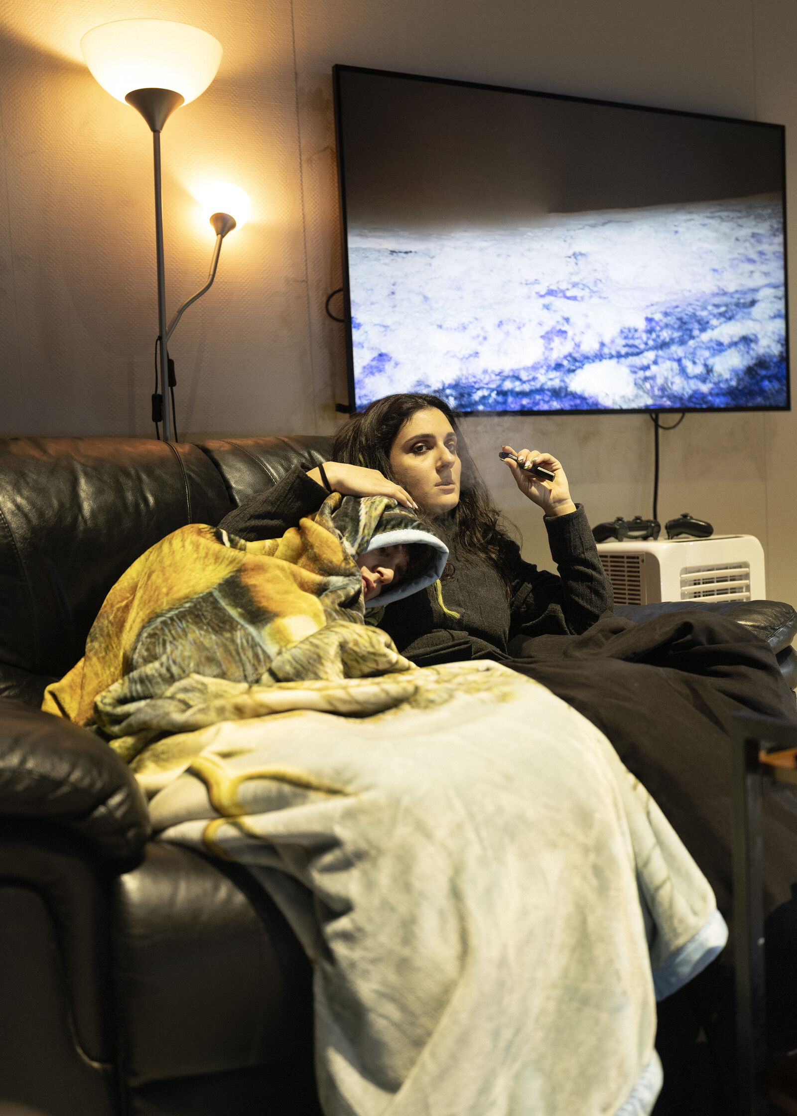 Zwei Personen liegen auf einem Sofa, bedeckt mit Decken. Eine lehnt an der anderen, die raucht. Im Hintergrund zeigt ein großer Bildschirm ein Landschaftsbild. Zwei Lampen sorgen für sanftes Licht im Raum. Daneben liegt ein Videospiel-Controller.