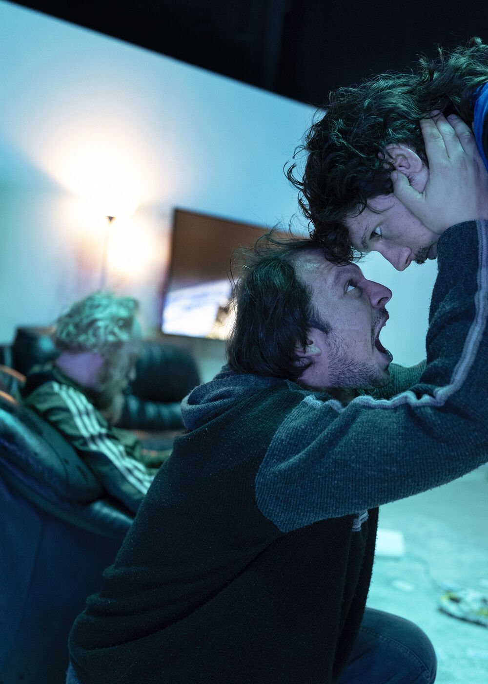 Eine dramatische Szene mit zwei Männern, die sich intensiv gegenüberstehen, wobei einer das Gesicht des anderen hält. Im Hintergrund sitzt eine Person auf einem Sofa. Die Szenerie ist von schwacher Beleuchtung und angespannter Atmosphäre geprägt.